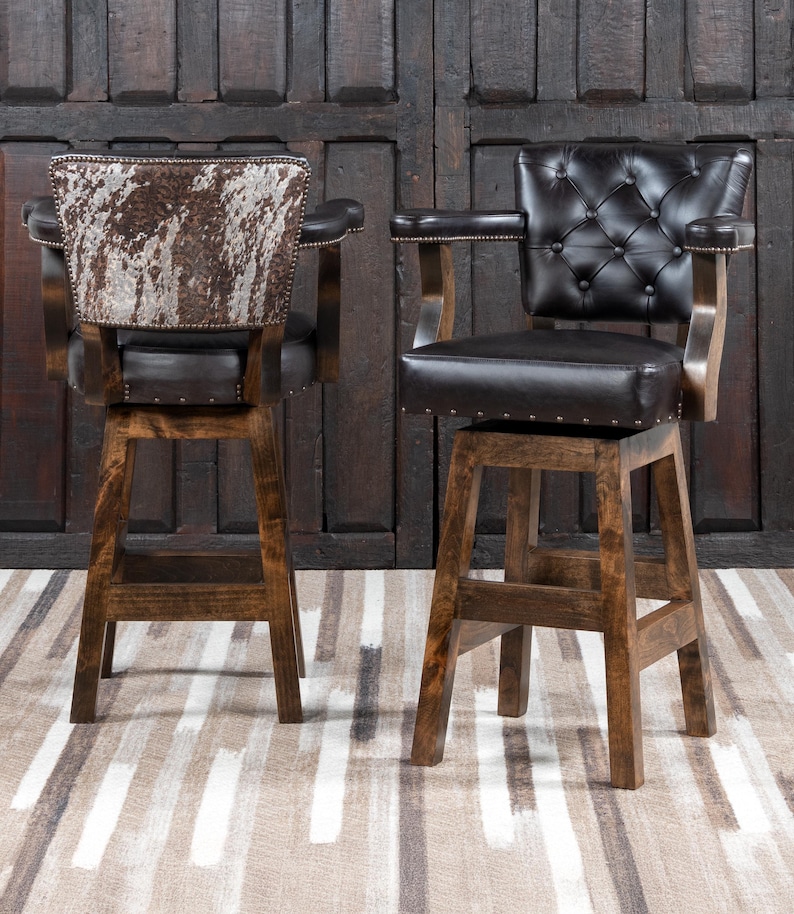 May include: Two wooden bar stools with dark brown leather seats and backs. One stool has a cowhide back, the other has a button-tufted back. Both have armrests and are on a light brown and white striped rug.