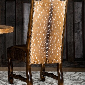 May include: A dark brown wooden chair with a high back featuring a spotted, tan and white hide. The seat is a darker brown leather. The chair has decorative nailhead trim and is set against a dark wood background.