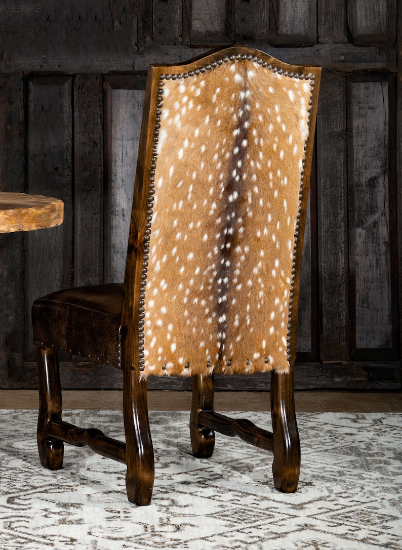 May include: A dark brown wooden chair with a high back featuring a spotted, tan and white hide. The seat is a darker brown leather. The chair has decorative nailhead trim and is set against a dark wood background.