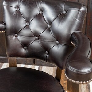 May include: Close-up of a brown leather chair with a tufted back and armrests. The chair has a dark brown leather seat and back, with button detailing. The armrests are curved and have a row of silver studs. The chair's wooden frame is visible.