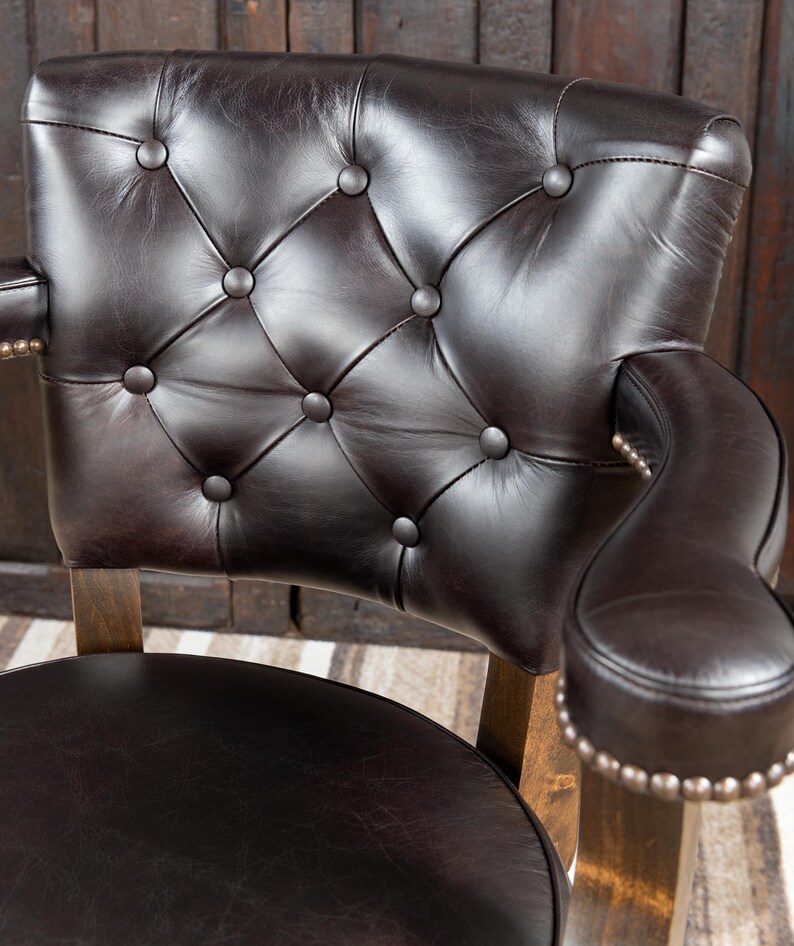 May include: Close-up of a brown leather chair with a tufted back and armrests. The chair has a dark brown leather seat and back, with button detailing. The armrests are curved and have a row of silver studs. The chair's wooden frame is visible.