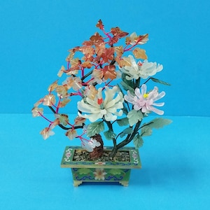 以下が含まれることがあります： 緑と青の塗装が施された鉢に、白、ピンク、茶色の花が咲いた翡翠の花の飾り。花は翡翠で作られており、鉢は花柄で飾られています。