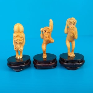 May include: Three small monkey figurines in various poses, each mounted on a dark brown, round wooden base. The monkeys are a light tan color, with detailed facial features. The background is a solid, bright blue.