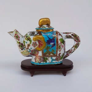 Pode incluir: Uma pequena chaleira de porcelana branca em forma de elefante, decorada com flores e pássaros de esmalte colorido. A chaleira tem uma tampa dourada e fica em um suporte de madeira.