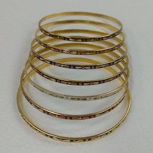 Può includere: Un set di otto braccialetti dorati con disegni in smalto colorati. I braccialetti sono impilati uno sopra l'altro, con il braccialetto inferiore che è il più grande e il braccialetto superiore che è il più piccolo. I braccialetti sono decorati con una varietà di colori, tra cui rosso, blu, verde e bianco.