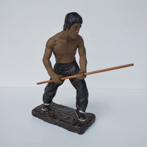 May include: A ceramic figurine of a martial arts figure holding a wooden staff. The figure is wearing black pants and has a bare chest. The figurine is standing on a brown base.