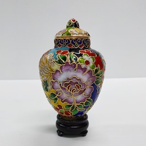 Pode incluir: Um pequeno vaso cloisonné colorido com um desenho floral. O vaso está sobre um suporte de madeira preto.