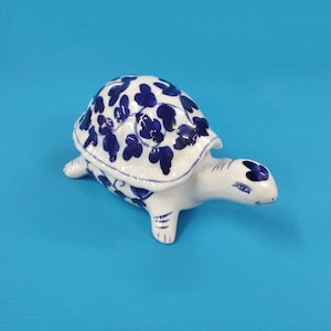 Könnte beinhalten: Eine weiße Keramik-Schildkrötenfigur mit einem blauen und weißen Blumenmuster. Die Schildkröte befindet sich in einer Gehposition mit ausgestrecktem Kopf.