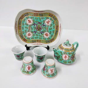 Peut inclure: Un service à thé en porcelaine vert et blanc avec un plateau, une théière et quatre tasses. L'ensemble présente un motif floral avec des caractères chinois.