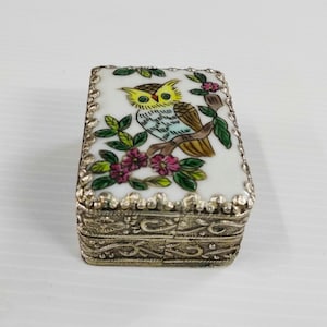 May include: A small, rectangular trinket box with a silver-colored metal base. The top features a white enamel design of a colorful owl perched on a branch with pink flowers and green leaves. The box is likely vintage.