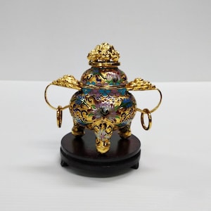 Peut inclure: Un petit brûleur d'encens de couleur or avec des motifs floraux complexes en émail bleu, rose et vert. Le brûleur repose sur une base en bois et a trois pieds.