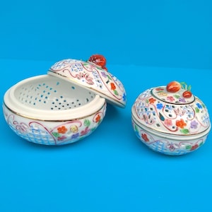 Puede incluir: Dos cajas de cerámica blanca para guardar pequeños objetos con intrincados diseños florales y detalles dorados. Las cajas están decoradas con flores rojas, azules y amarillas y tienen un patrón de celosía. La tapa de una caja está abierta, revelando el interior vacío.