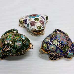 May include: Three cloisonné frog figurines with intricate floral designs. The frogs are painted in green, white, and blue with gold accents.