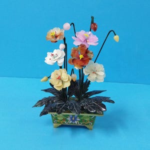 Op de afbeelding: Een klein, decoratief bloemstuk gemaakt van gesneden jade en andere edelstenen. De bloemen staan in een groene en gouden pot met een bloemenmotief.