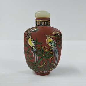 May include: A small, red, ceramic snuff bottle with a white jade stopper. The bottle is decorated with two birds perched on a branch with green leaves and yellow flowers.