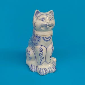 Puede incluir: Una figura de gato de cerámica blanca con diseños decorativos azules. El gato está sentado con las orejas levantadas. Los diseños azules incluyen motivos florales y de remolino, creando una apariencia detallada y artística. El fondo es azul liso.
