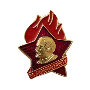 Könnte beinhalten: Eine rote Emaille-Anstecknadel mit einem goldenen Stern und einem Porträt eines Mannes. Der Stern hat oben ein Flammenmotiv und unten ein rotes Band mit dem Text "ЗА АКТИВНУЮ РАБОТУ".