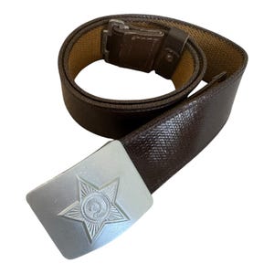 Op de afbeelding: Een donkerbruine, getextureerde riem met een rechthoekige, zilverkleurige metalen gesp. De gesp toont een in reliëf gemaakte vijfpuntige ster met een hamer en sikkel embleem in het midden. De riem heeft een beige voering aan de binnenzijde.