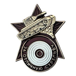 May include: A military-style pin featuring a tank on a star-shaped emblem. The pin has a gold-colored tank and a dark red star with white and gold accents. The text on the pin is in Russian.