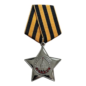 Può includere: Una medaglia a forma di stella a cinque punte in argento con una scritta in smalto rosso che recita "СЛАВА" (Gloria) e una rappresentazione di un edificio con una stella rossa in cima. La medaglia è attaccata a un nastro a strisce nere e gialle.
