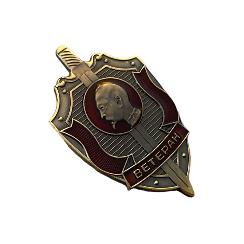 Pode incluir: Um broche de metal dourado em forma de escudo com um design vermelho e dourado. O escudo apresenta um perfil de uma pessoa com uma espada e a palavra "Ветеран" em escrita cir&iacute;lica.