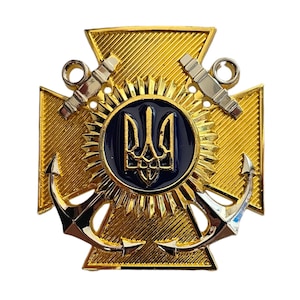 May include: A gold-colored military badge with a cross design. The badge features a central blue emblem with a gold trident, surrounded by a sunburst pattern. Two silver cannons and two anchors are also present.