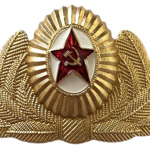 May include: A gold-colored military cap badge with a red star and hammer and sickle emblem. The star is set against a white background and the badge is surrounded by a wreath of gold leaves.