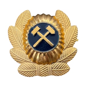 May include: A gold-colored emblem featuring a central blue circle with crossed hammers. The emblem is surrounded by a gold sunburst design and detailed leaf-like elements, creating a decorative and ornate appearance.