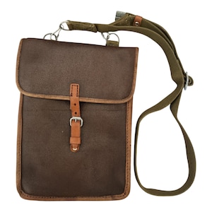 May include: A brown leather messenger bag with a flap closure and a tan leather strap. The bag has a green canvas shoulder strap and metal hardware. The edges of the bag are trimmed with tan leather.
