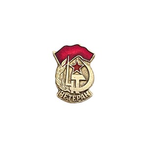 Puede incluir: Un pin de color dorado con un diseño de estrella roja, martillo y hoz. El pin tiene la palabra "Veterano" escrita en cirílico.