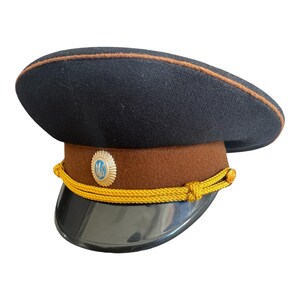 Könnte beinhalten: Eine dunkelblaue Militärmütze mit braunem Band und schwarzem Visier. Die Mütze hat goldfarbene Seildetails und ein rundes Emblem. Der Rand ist mit einer braunen Zierleiste versehen.