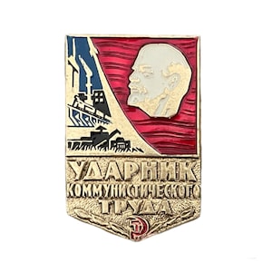 Puede incluir: Un pin de metal dorado con un diseño rojo, blanco y azul. El pin presenta un perfil de un hombre con barba y una escena de fábrica. El texto "УДАРНИК КОММУНИСТИЧЕСКОГО ТРУДА" está escrito en el pin.