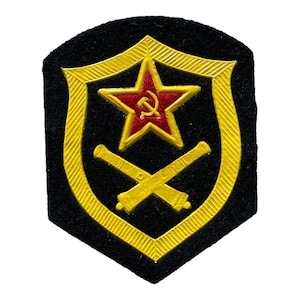 May include: A black patch featuring a gold shield design with a red star and hammer and sickle emblem at the center. Two crossed cannons are below the star. The patch has a gold border.