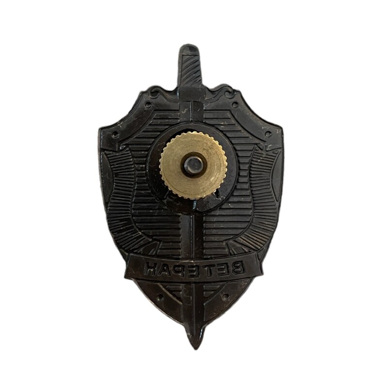 Pode incluir: Um distintivo de metal preto em forma de escudo com uma espada e a palavra "ВЕТЕРАН" em escrita cir&iacute;lica. O distintivo tem um pino dourado na parte de tr&aacute;s.