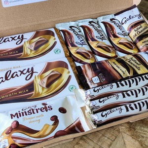 May include: A brown cardboard box filled with various chocolate bars and bags. The box contains Galaxy Smooth Milk chocolate bars, Galaxy Minstrels, Galaxy Ripple chocolate bars, and Galaxy Instant Hot Chocolate.