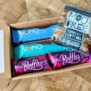 May include: An open cardboard box filled with various chocolate bars. The box is decorated with a birthday hat and the word "sage". The chocolate bars include two blue and white "NOMO" bars, two purple "Ruffles" bars, and a bag of "Moo Free Choccy Buttons" for dairy dodging.