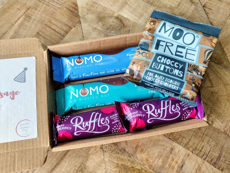May include: An open cardboard box filled with various chocolate bars. The box is decorated with a birthday hat and the word "sage". The chocolate bars include two blue and white "NOMO" bars, two purple "Ruffles" bars, and a bag of "Moo Free Choccy Buttons" for dairy dodging.