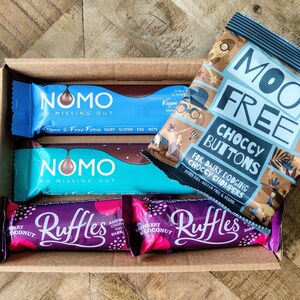 May include: A box filled with various chocolate bars and a bag of chocolate buttons. The chocolate bars are wrapped in blue, purple, and pink foil. The bag of chocolate buttons is blue and brown with the text "Moo Free Choccy Buttons For Dairy Dodging Choccy Chompers Dairy Free Gluten Free & Vegan".
