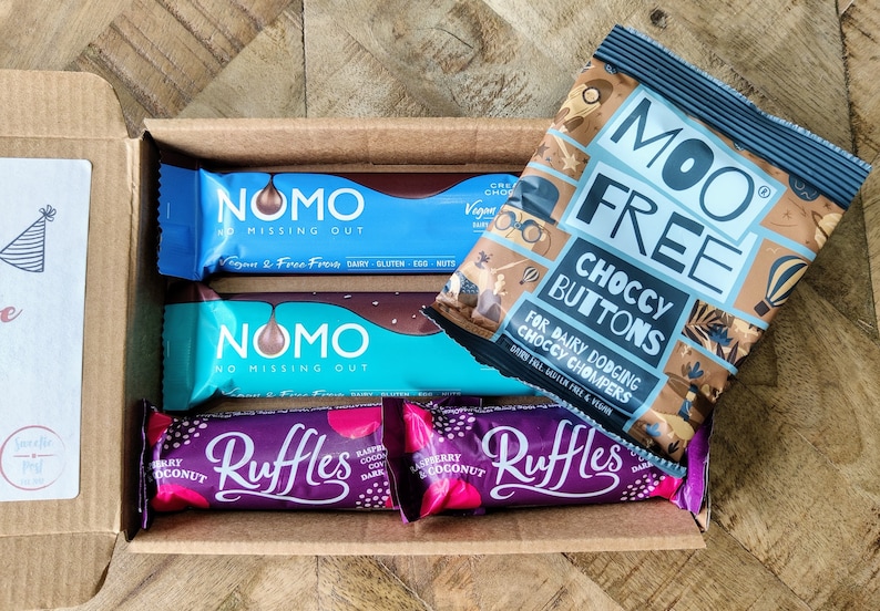May include: A box filled with various chocolate bars and a bag of chocolate buttons. The chocolate bars are wrapped in blue, purple, and pink foil. The bag of chocolate buttons is blue and brown with the text "Moo Free Choccy Buttons For Dairy Dodging Choccy Chompers Dairy Free Gluten Free & Vegan".