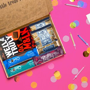 May include: An open cardboard box filled with various chocolate bars and snacks. The box is on a pink surface with confetti and birthday candles. The snacks include Well & Truly, Oat M&LK, Moo Free Choccy Buttons, Nomo, and Ruffles.
