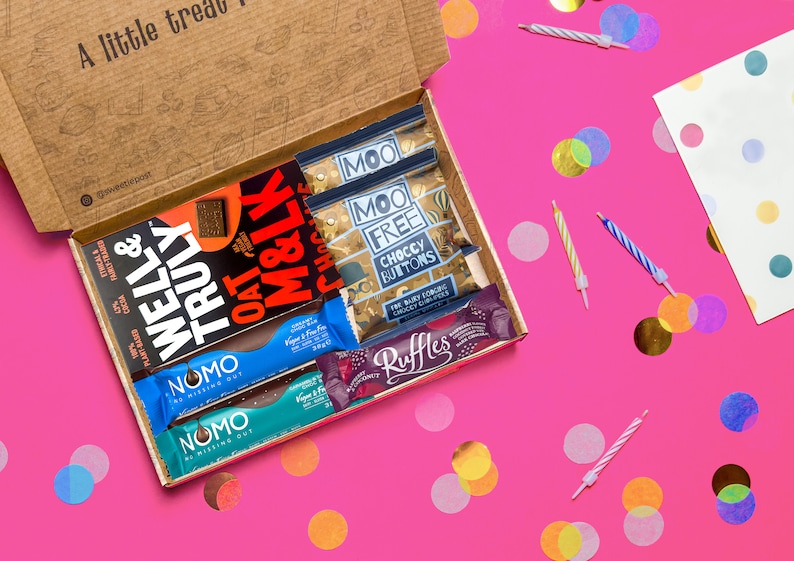 May include: An open cardboard box filled with various chocolate bars and snacks. The box is on a pink surface with confetti and birthday candles. The snacks include Well & Truly, Oat M&LK, Moo Free Choccy Buttons, Nomo, and Ruffles.