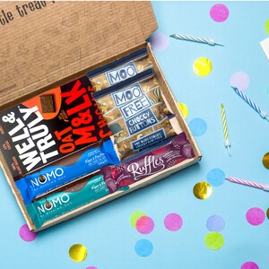 May include: A brown cardboard box filled with various chocolate bars and snacks. The box is open and the contents are visible. The snacks include a chocolate bar with the words "Well & Truly" on it, a chocolate bar with the words "Oat & Milk" on it, a chocolate bar with the words "Moo Free Choccy Buttons" on it, and a chocolate bar with the words "Ruffles" on it. The box is sitting on a blue surface with colorful confetti scattered around it.