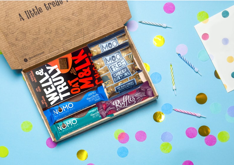 May include: A brown cardboard box filled with various chocolate bars and snacks. The box is open and the contents are visible. The snacks include a chocolate bar with the words "Well & Truly" on it, a chocolate bar with the words "Oat & Milk" on it, a chocolate bar with the words "Moo Free Choccy Buttons" on it, and a chocolate bar with the words "Ruffles" on it. The box is sitting on a blue surface with colorful confetti scattered around it.