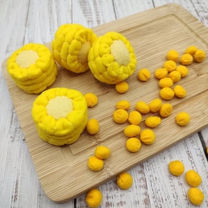 Puede incluir: Cuatro maíces de fieltro amarillo en una tabla de cortar de madera con granos de maíz de fieltro amarillo esparcidos alrededor.