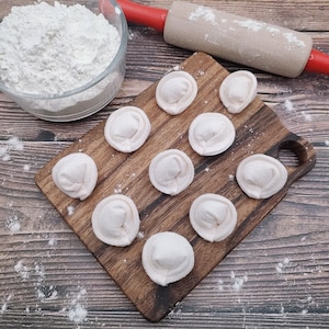 May include: A wooden cutting board with nine white dumplings arranged in a circular pattern. The board is on a wooden surface with flour scattered around it.