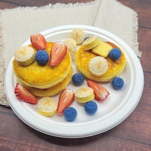 May include: A white plate with a stack of three yellow pancakes topped with strawberries, blueberries, and banana slices. The pancakes are surrounded by additional fruit and blue and yellow decorative balls.