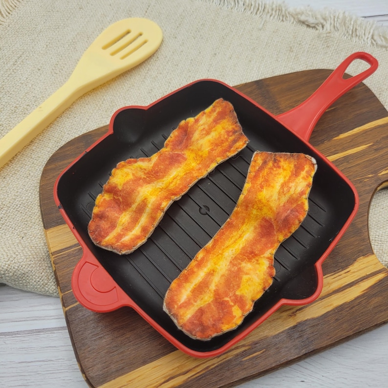 Op de afbeelding: Twee plakjes rood en geel gekleurd spek liggen in een zwarte en rode vierkante grillpan. Een gele sleuflepel bevindt zich links van de pan. De pan staat op een houten snijplank.