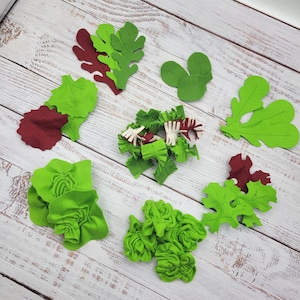 May include: An overhead shot displays various felt food pieces, including green and maroon lettuce leaves, and chopped lettuce. The pieces are arranged on a light wood-grain surface, suggesting a craft or play food set.