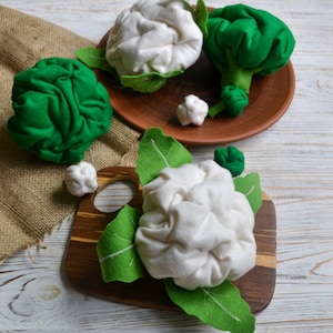 May include: Three felt cauliflower heads with green felt leaves on a wooden cutting board and a brown plate. The cauliflower heads are white and the leaves are green.