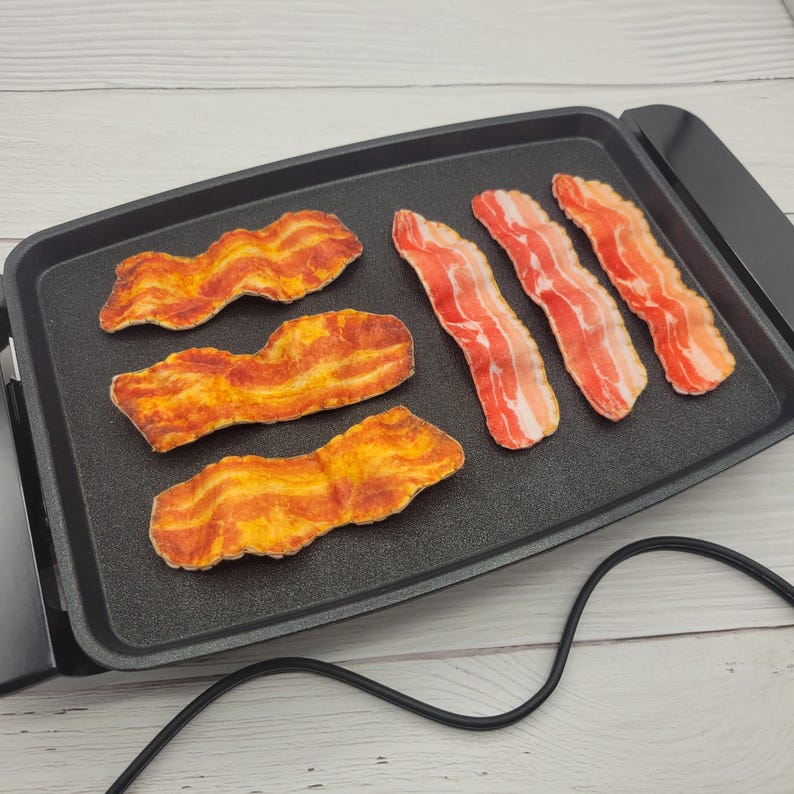 Op de afbeelding: Een zwarte elektrische grill met zes plakjes gebakken spek. Het spek is een mix van rood en geel, met een knapperige uitstraling. De grill heeft een antiaanbakoppervlak en een zwart netsnoer.